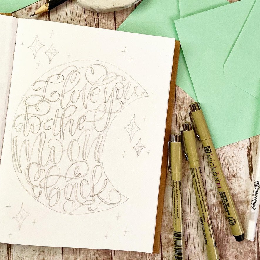 a pencil sketch in a notebook of hand lettering in a moon shape